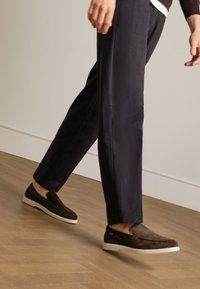 Brown suede slip-on shoes with a white sole, paired with navy trousers, captured mid-stride on a wooden floor.