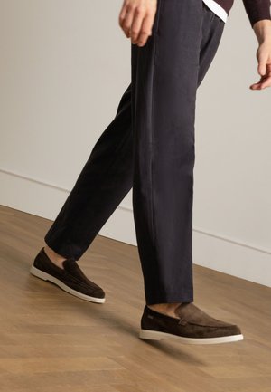 Des chaussures en suede marron à enfiler avec une semelle blanche, associées à un pantalon marine, capturées en plein mouvement sur un parquet en bois.