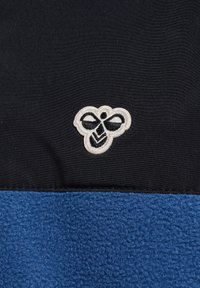 Zwart met blauw fleece kledingstuk met wit geborduurd bijenlogo. Heeft een glad bovenste gedeelte en een getextureerd blauw onderste gedeelte.