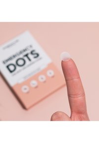 Patch circulaire transparent reposant sur un bout de doigt, avec une texture douce ; un emballage rose en partie visible étiqueté "Emergency Dots" à l'arrière-plan.