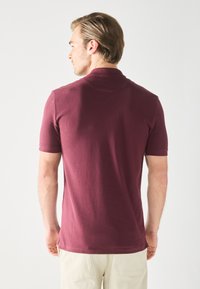 Maronfarvet polo-shirt med korte ærmer, glat tekstur og et diskret kravedesign. Bagsiden viser en tætsiddende silhuet og rene linjer.