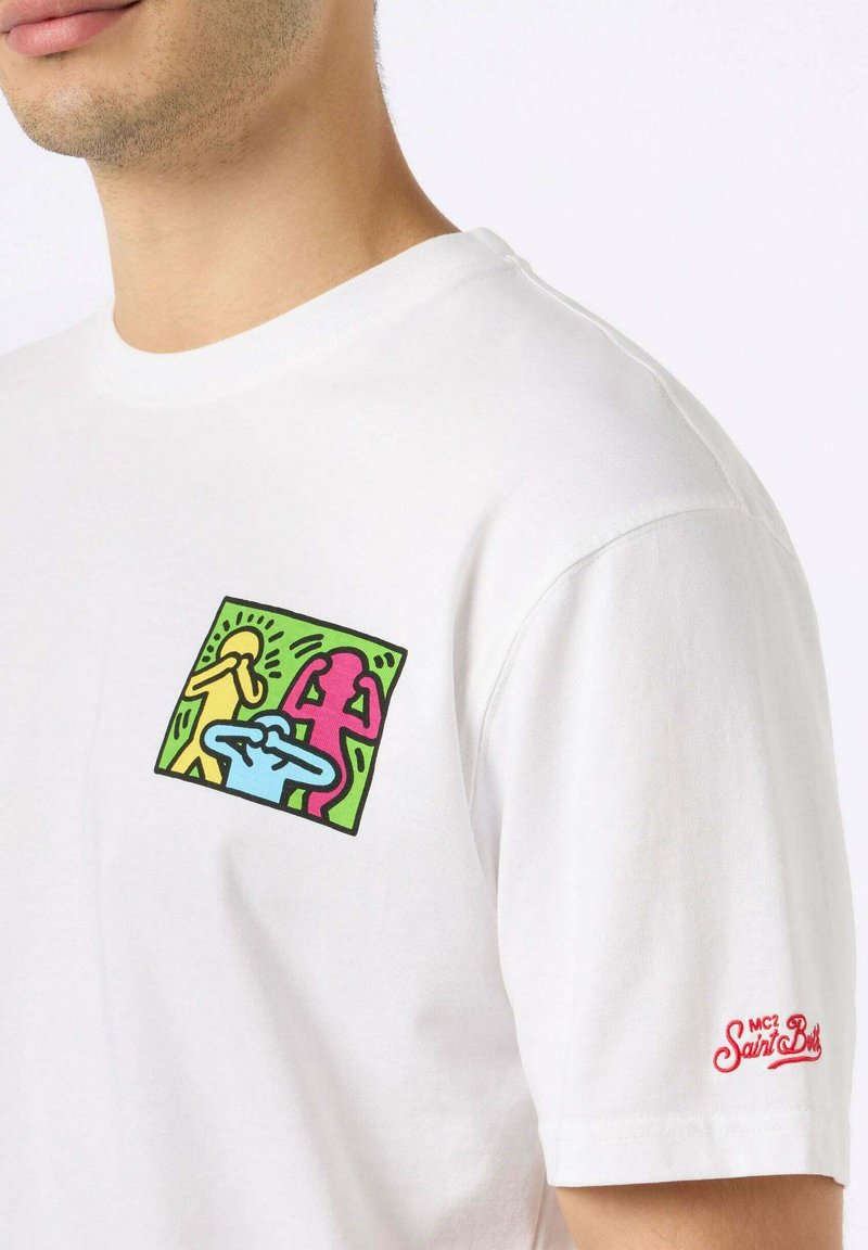 T-shirt bianco in cotone con una patch grafica colorata che presenta figure astratte in giallo, rosa e blu sul lato sinistro del petto, logo rosso sulla manica.