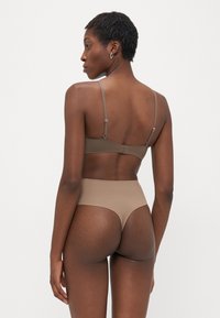 Woman with short hair wearing taupe bra and high-waist thong underwear, standing with back facing camera on plain white background.