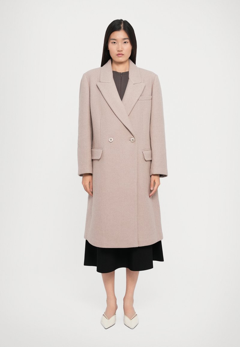 Woman wearing a long beige double-breasted coat over a black skirt and white pointed-toe shoes, standing against a plain white background.