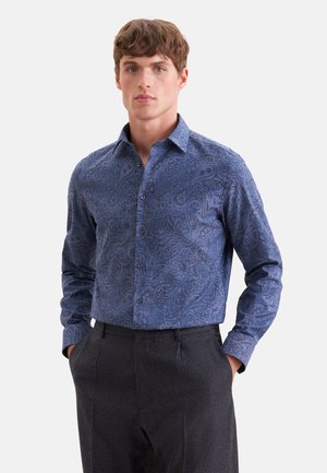 Giovane uomo che indossa una camicia blu a bottoni con motivo paisley e pantaloni grigio scuro, con le mani nelle tasche, in piedi davanti a uno sfondo semplice.