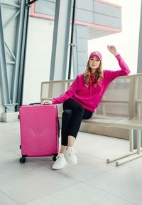 Růžový tvrdý kufr s embosovaným vzorem vln, čtyřmi kolečky a teleskopickou rukojetí, umístěný vedle jednotlivce v růžové mikině s kapucí a čepici.