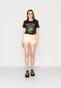 Camiseta preta cropped com texto gráfico, combinada com calções tie-dye laranja claro. Modelo usa sandálias pretas de plataforma e óculos de sol amarelos.