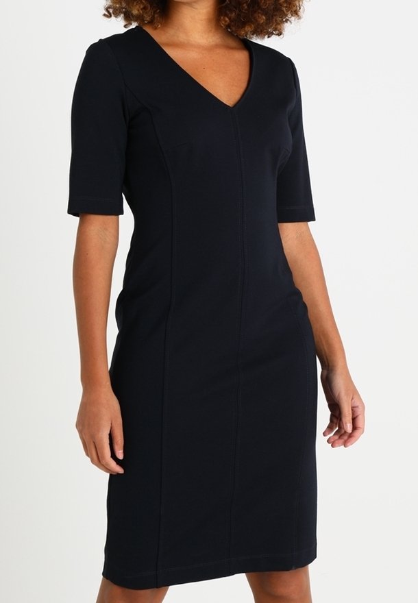 Femme portant une robe noire ajustée longueur genou avec manches aux coudes et décolleté en V, debout sur un fond blanc uni.