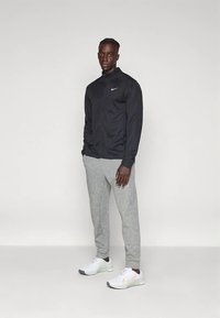 Černá atletická bunda na zip s logem Nike, v kombinaci se světle šedými tepláky a bílými teniskami s oranžovým detailem.