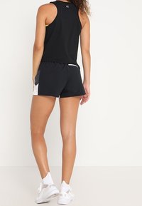 Sort ærmeløs top med en gul detalje, parret med sorte shorts med hvide sidepaneler; båret med hvide træningssko.