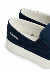 Le scarpe slip-on in tela blu navy presentano una suola in gomma bianca, un design con cuciture e un piccolo etichetta di marca con accenti rossi e blu.