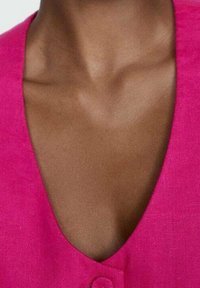 Top fuchsia à col en V avec un tissu texturé et un détail de bouton unique au niveau de l'encolure, contrastant avec un teint de peau lisse.