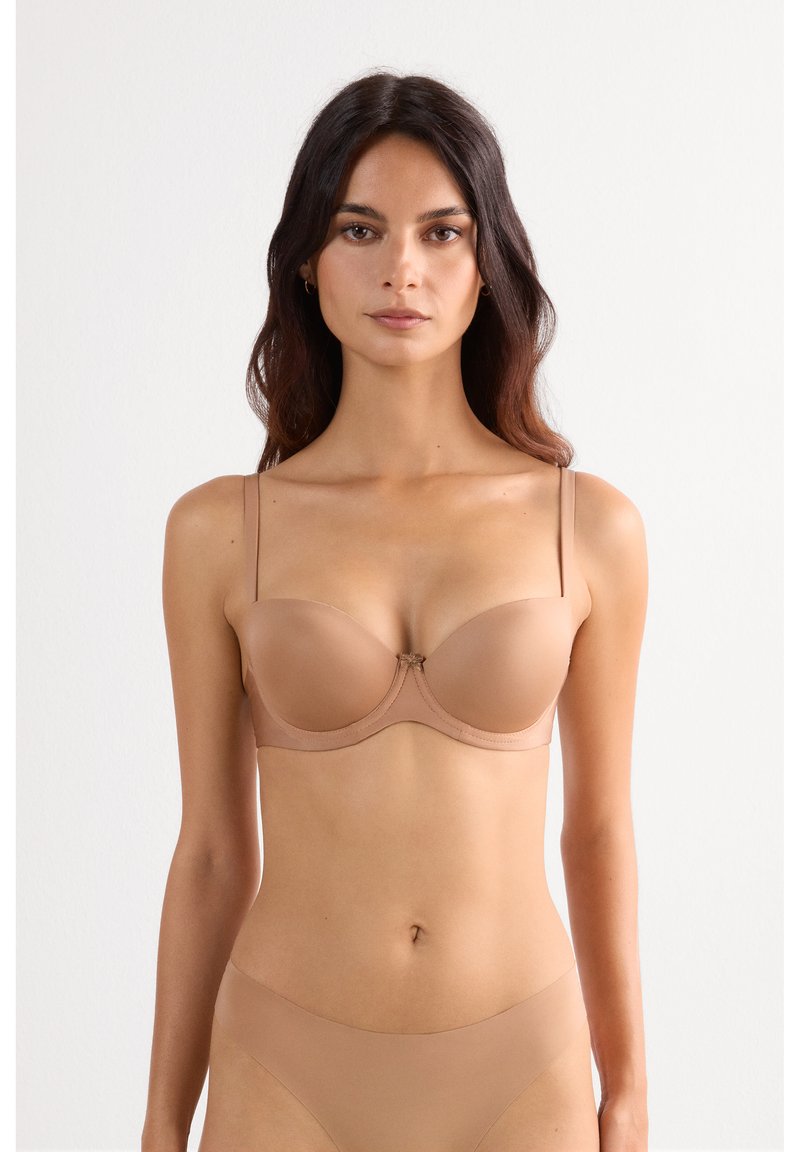 Kvinde med langt mørkt hår iført en nude-farvet strapless bh og matchende sømløs undertøj mod en ensfarvet hvid baggrund.