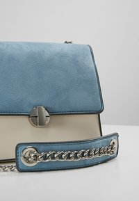 Světle modrá semišová kabelka s překlopeným klopou, krémovým tělem, stříbrnými detaily a ramenním popruhem s řetězovou dekorací. Hladká textura, moderní design.