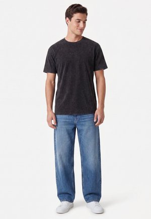 Giovane uomo che indossa una maglietta nera sbiadita, jeans blu larghi e scarpe da ginnastica bianche, in piedi davanti a uno sfondo bianco semplice.