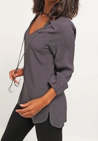 Langärmelige graue Bluse mit V-Ausschnitt und hochgekrempelten Ärmeln; verfügt über eine Kordelzug-Akzentuierung und einen leicht gebogenen Saum. Glatte Textur.