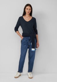 Marineblaues Langarm-V-Ausschnitt-Oberteil kombiniert mit dunkelblauen, hoch taillierten Jeans mit hellblauen Akzenten und weißen Sneakers mit beigefarbenen Sohlen.