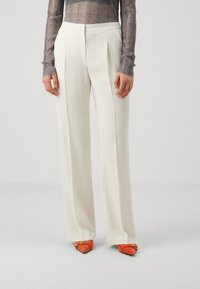 Pantalons plissés blancs à coupe ajustée, fabriqués en tissu lisse. Associés à des chaussures à bouts pointus orange avec des éléments décoratifs.