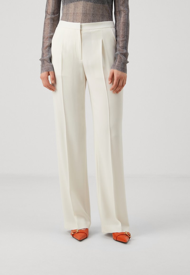 Pantalons plissés blancs à coupe ajustée, fabriqués en tissu lisse. Associés à des chaussures à bouts pointus orange avec des éléments décoratifs.