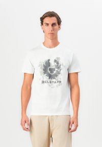 Balta medvilninė marškinėliai su pilkai juoda grafika, kuriame yra apvalus emblema ir tekstas "BELSTAFF" apačioje, trumpomis rankovėmis.