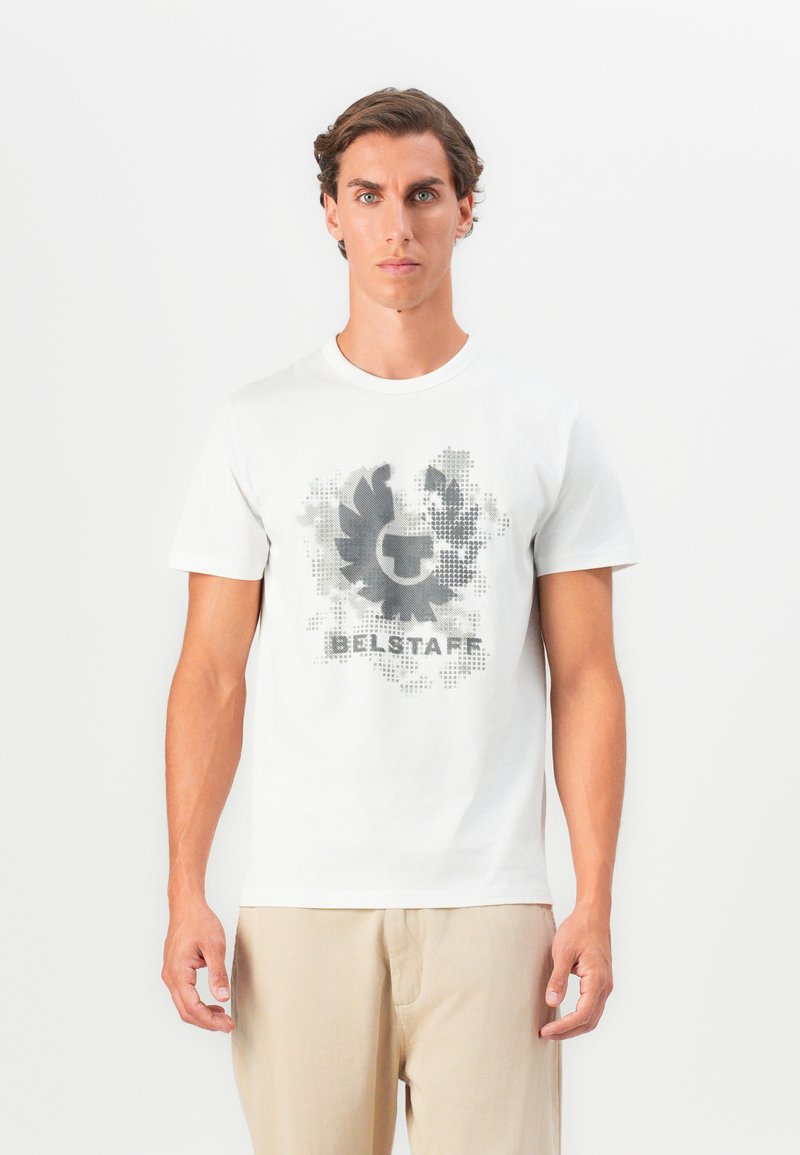 Balta medvilninė marškinėliai su pilkai juoda grafika, kuriame yra apvalus emblema ir tekstas "BELSTAFF" apačioje, trumpomis rankovėmis.