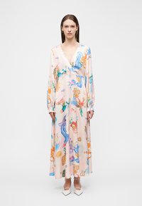 Γυναίκα στέκεται μπροστά φοράει μακρύ παστέλ φόρεμα με πολύχρωμο θέμα υποβρύχιας ζωής και λευκά μυτερά τακούνια.