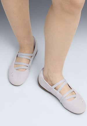 Füße in hellgrauen Ballerinas mit drei elastischen Riemen über dem Spann, stehend auf einem grauen Verlaufshintergrund.