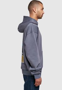 Szürke kapucnis sweatshirt, bő szabással, nagy elülső zsebbel és színes grafikai nyomattal a háton. Puha anyag, bordázott mandzsetták.