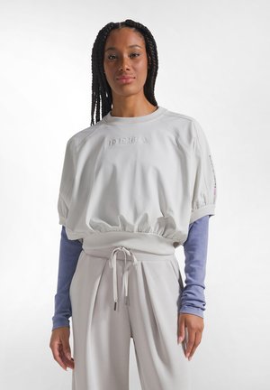 Vrouw met gevlochten haar die een losse witte cropped sweatshirt draagt over een lavendelkleurige top met lange mouwen en bijpassende witte broek met koord.