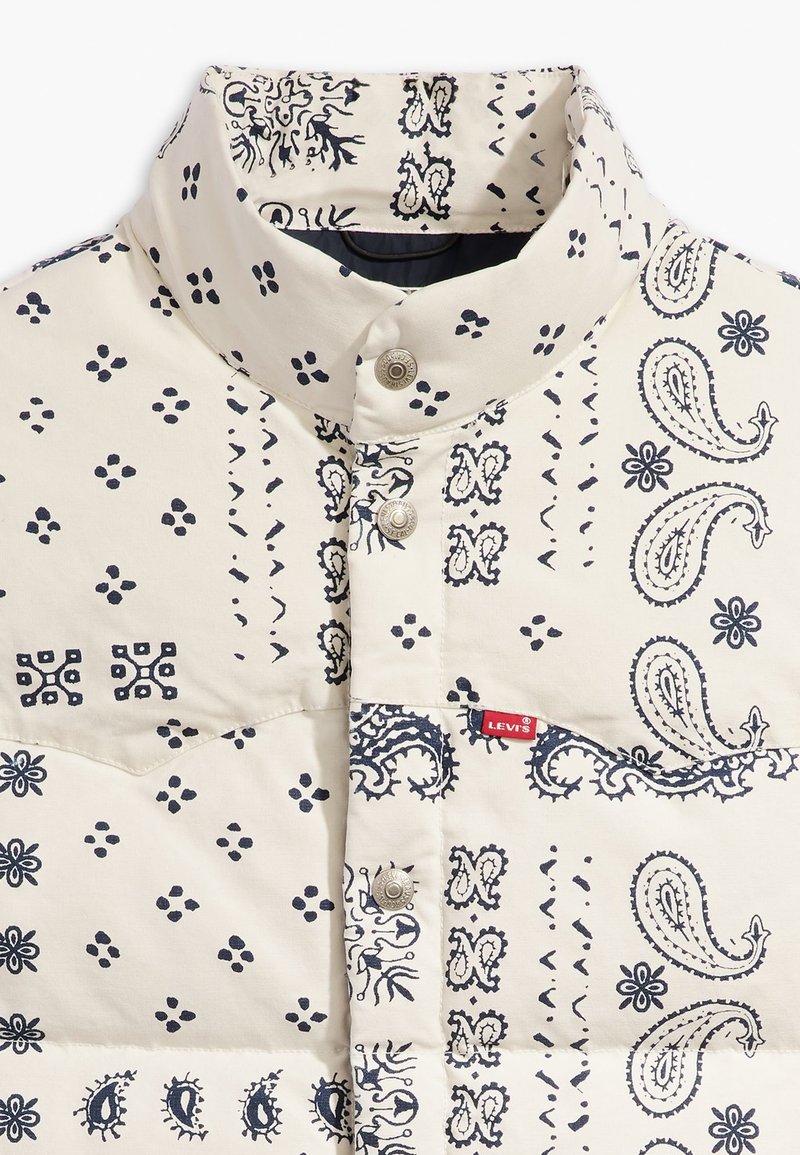 Chaqueta acolchada de tela crema con patrones de paisley y flores en azul marino. Presenta un cuello alto y botones a presión, con una etiqueta de marca.