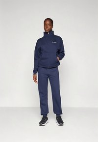 Frau, die in einem marineblauen Trainingsanzug mit Reißverschlussjacke und schwarzen Sportschuhen vor einem schlichten hellgrauen Hintergrund steht.