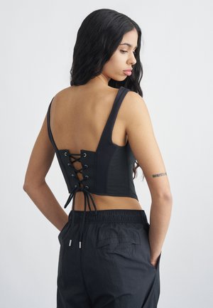 Kvinne med langt svart hår iført en svart crop top med snøring bak og svarte bukser, stående med hendene i lommene, vendt sidelengs.