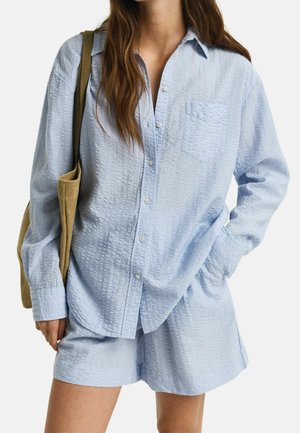 Kvinde iført lyseblå tekstureret skjorte med knapper og matchende shorts med en beige skuldertaske.
