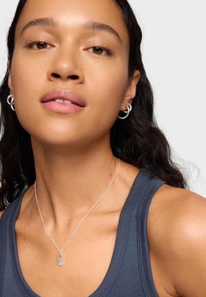 Kobieta nosząca srebrne kolczyki koła i srebrny łańcuszek z owalnym zawieszeniem, ubrana w ciemny, bezrękawnik na jednolitym tle.