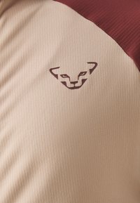 Beige Sportshirt mit einem burgunderfarbenen Schulterakzent; verfügt über einen strukturierten Stoff und ein erhabenes Logo eines stilisierten Tiergesichts.