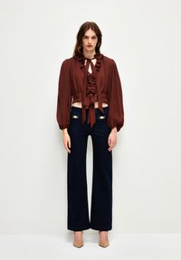 Blusa recortada de color marrón rústico con volantes y detalle de lazo. Combinada con pantalones de pierna ancha en color azul marino con acentos en dorado. Tacones neutros.