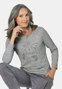 Grau Sweater mit langen Ärmeln, rundem Halsausschnitt und einem geometrischen Muster aus Strasssteinen auf der Vorderseite. Kombiniert mit grauen Wildlederhosen.