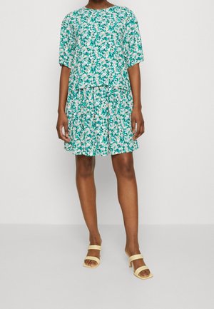 Vrouw draagt een loszittende, korte mouwen jurk met bloemenprint in groen en blauw, gecombineerd met crèmekleurige sandalen met hak.