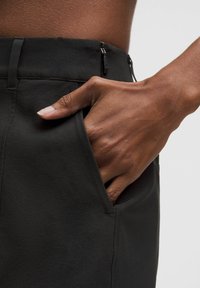 Pantalones negros con una textura suave y un ajuste entallado. Cuentan con bolsillos laterales y un cierre de cremallera oculto. Primer plano de la mano en el detalle del bolsillo.