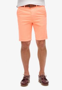 Pantaloni corti leggeri, di lunghezza al ginocchio, in tessuto corallo brillante, con vita piatta, tasche laterali e texture liscia. Abbinati a scarpe da barca marroni.