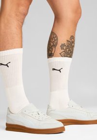 Sneakers bianchi con suola in gomma, caratterizzati da un design elegante, pelle testurizzata e perforazioni, abbinati a calzini bianchi a coste e logo Puma.