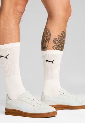 Baskets blanches avec une semelle en caoutchouc, présentant un design épuré, un cuir texturé et des perforations, associées à des chaussettes blanches côtelées avec le logo Puma.