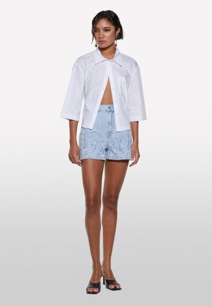 Chemise blanche à manches courtes et col, associée à un short en denim bleu avec des broderies violettes et taille haute.