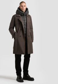 Brown trench coat with a detachable puffer hooded layer, double-breasted buttons, and black pants; worn with black lace-up boots.