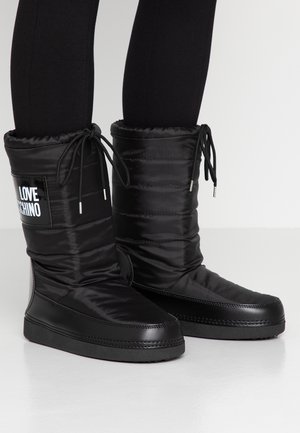 Musta keskipitkäpäällysteiset saappaat, joissa on kiristysnyörit ja "LOVE MOSCHINO" -logotarrat, pukeutuneena mustujen leggingsien päälle valkoisella lattialla.