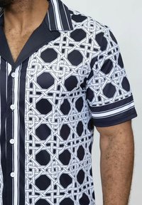 Chemise à manches courtes avec un motif géométrique noir et blanc, des motifs entrelacés et des rayures verticales contrastées au niveau du col et des poignets.