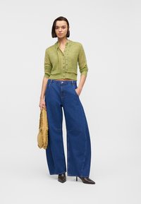 Femme portant un cardigan court vert boutonné, un jean bleu à jambes larges, des bottes noires à talons, tenant un sac marron à franges, debout devant un fond blanc.