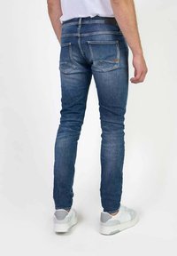 Jeans de mezclilla azul oscuro con un corte ajustado, que presentan desvaneos sutiles, bolsillos traseros y una cinturilla negra, combinados con zapatillas blancas.