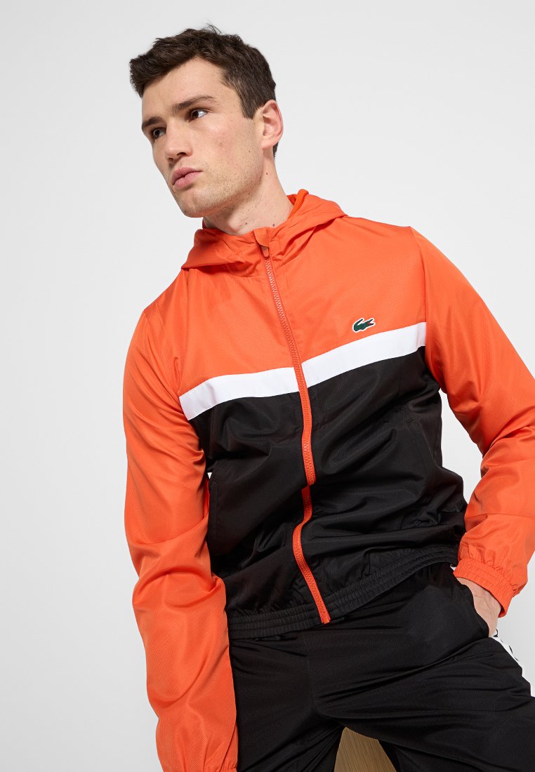 Veste coupe-vent pour homme avec une partie supérieure orange vif, une bande blanche, une partie inférieure noire et une capuche, fabriquée en matériau léger.