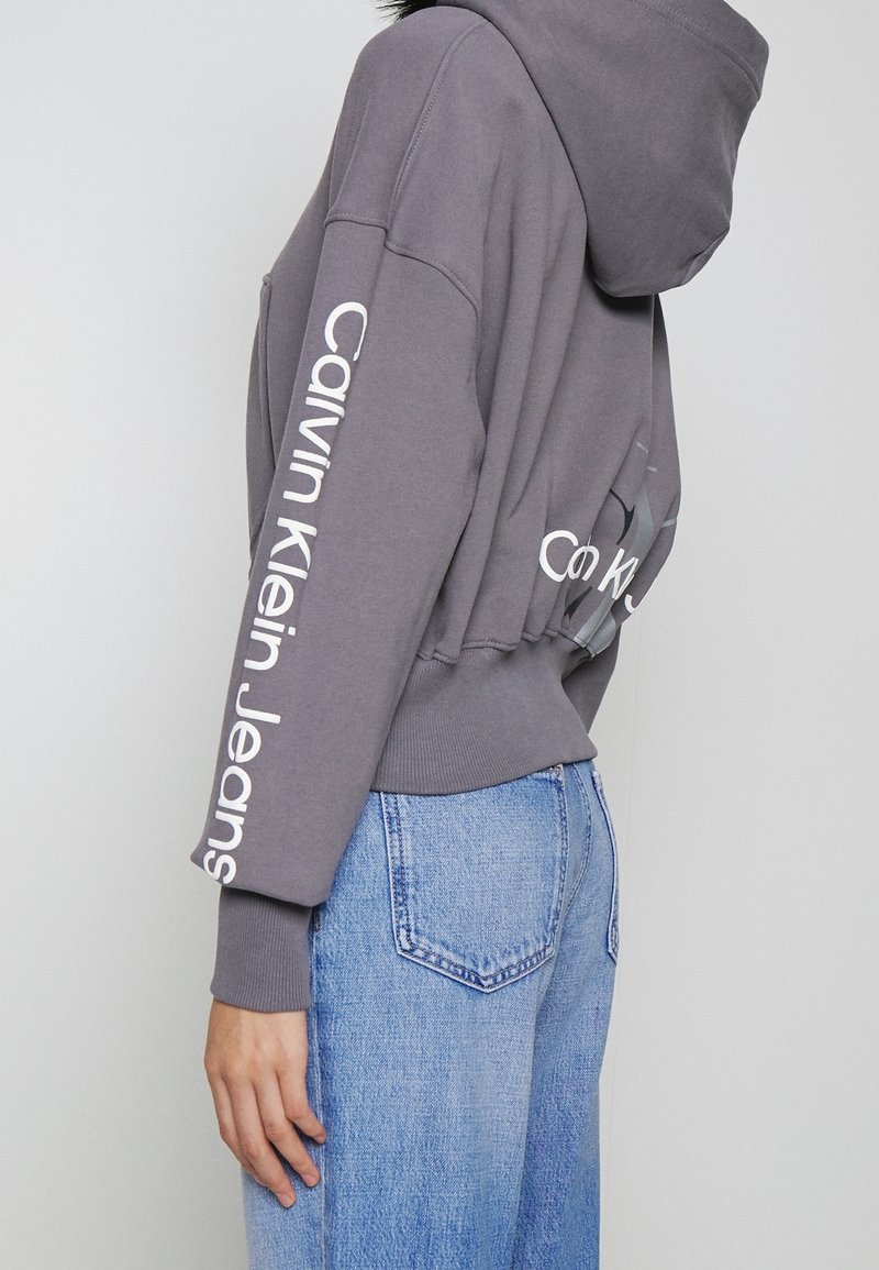 Persoon die een cropped grijze Calvin Klein Jeans hoodie en lichtblauwe high-waisted denim jeans draagt, zijaanzicht.
